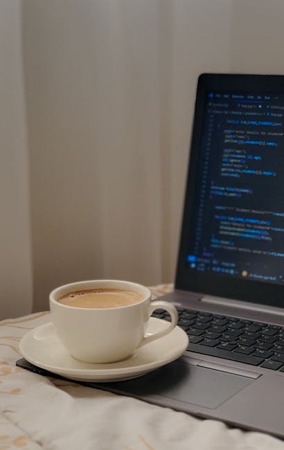 A young woman with shoulder-length light brown hair and fair skin is sitting at a dark table outdoors, working on a silver laptop. She is wearing a white shirt and has a blue and white striped scarf d